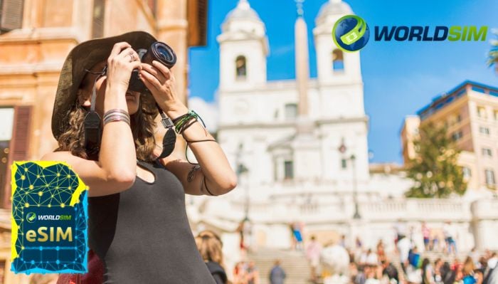 A tourist with a camera stands in front of a historic building, capturing the moment, with the WorldSIM logo and an image of a WorldSIM eSIM card visible