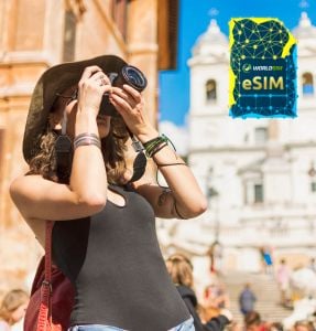 A tourist with a camera stands in front of a historic building, capturing the moment, with the WorldSIM logo and an image of a WorldSIM eSIM card visible