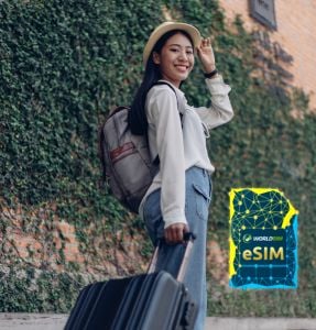 A woman traveler with a suitcase stands in front of a brick wall with greenery, smiling and looking back, with the WorldSIM logo and an image of a WorldSIM eSIM card shown